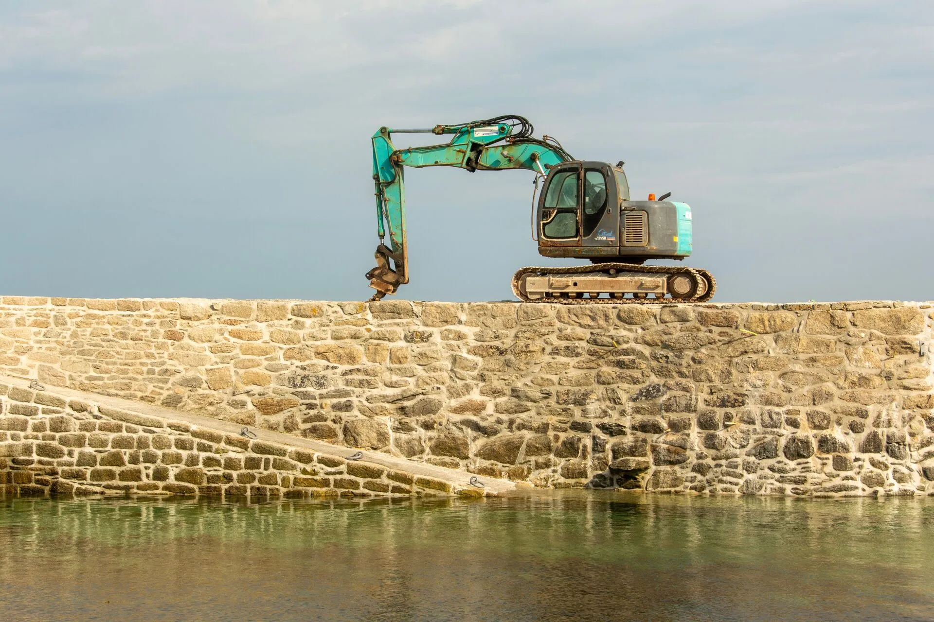 A construction excavator positioned on a stone pier by calm water